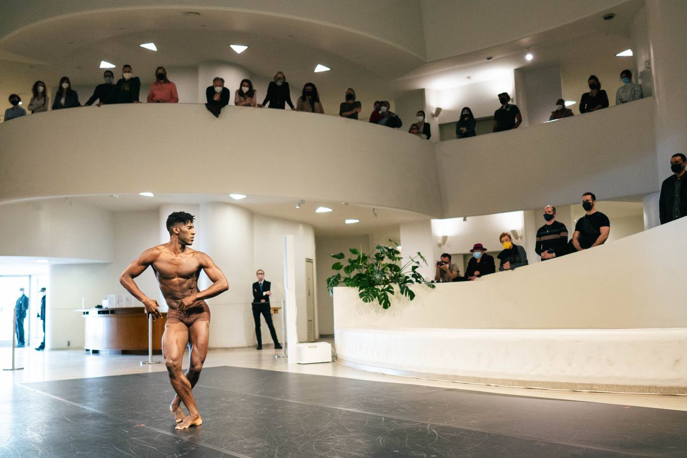 Attired in skin-toned briefs, Lloyd Knight spirals his body as he crosses his feet; people watch from above